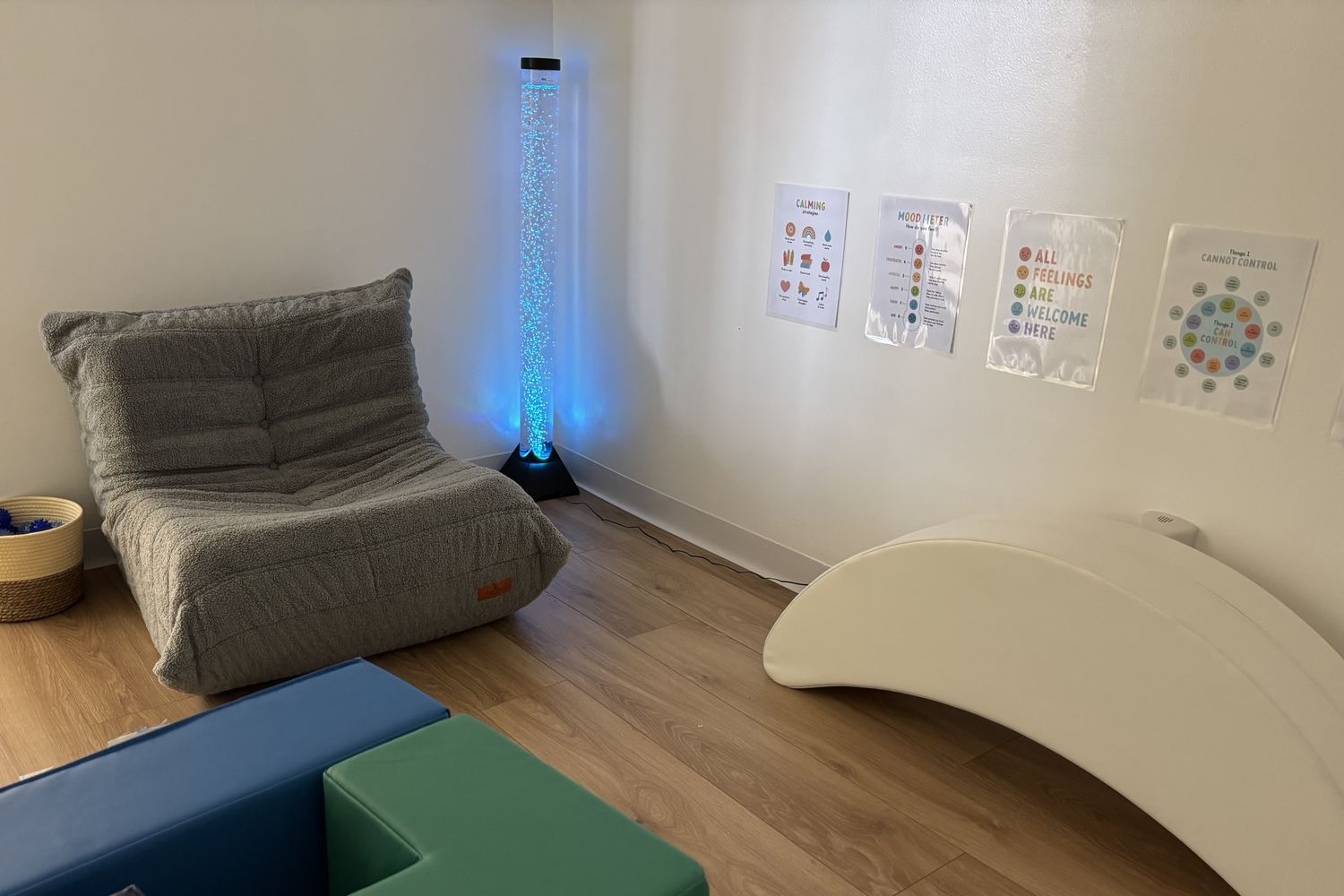 The mindfulness room: a glowing blue bubble tube in the corner beside a grey bean-bag lounger, a white wave rocker along the far wall, soft foam blocks on the floor and calming cards hung at child height.